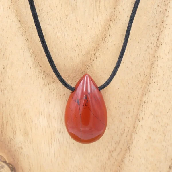 Pendentif en forme de goutte en jaspe rouge poli avec des veines noires naturelles, monté sur un cordon fin en coton ciré noir et présenté sur un fond en bois texturé.